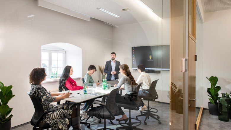 Stift Klosterneuburg, Quarter 1114, © GAS A modern conference room with six people sitting at a table listening to a speaker. A screen in the background.