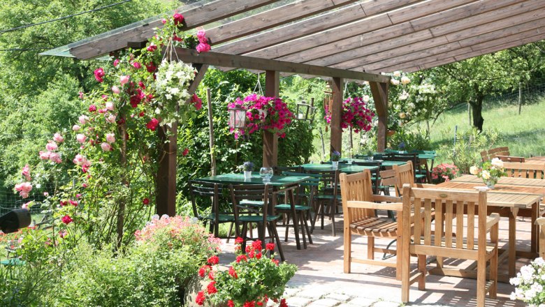 Ein gemütlicher Außenbereich eines Heurigen mit Holztischen und -stühlen, umgeben von blühenden Pflanzen und einem hölzernen Pergola-Dach.