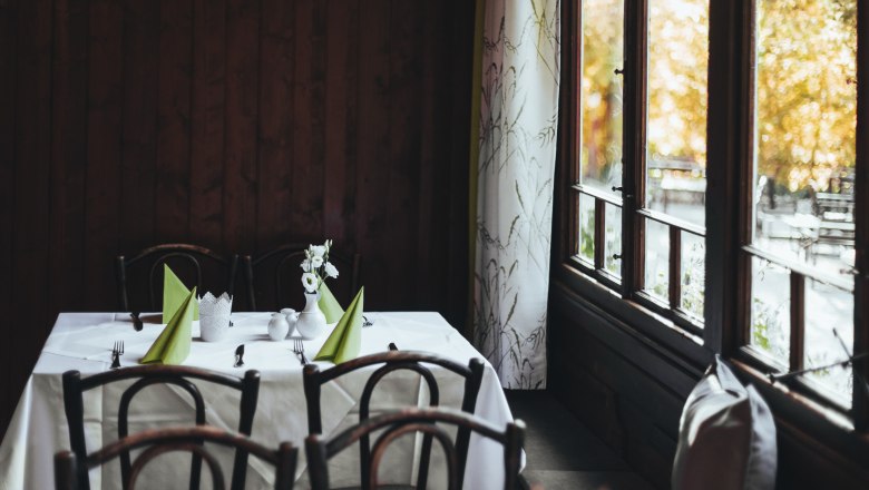 Ein Tisch in einer Gaststube mit Holzst&uuml;hlen, wei&szlig;en Tischdecken und gr&uuml;nen Servietten, neben einem Fenster mit Vorh&auml;ngen.