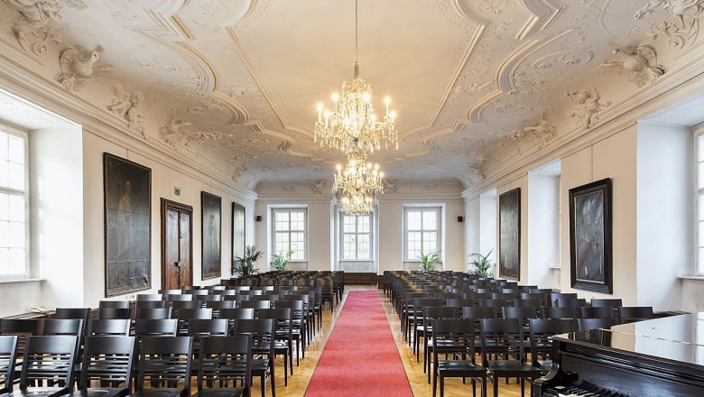 Elegant hall with stucco ceiling, chandeliers, rows of chairs and red carpet.