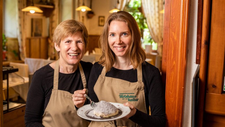 Zwei Frauen in Schürzen halten einen Teller mit einem Dessert in einem gemütlichen Restaurant.