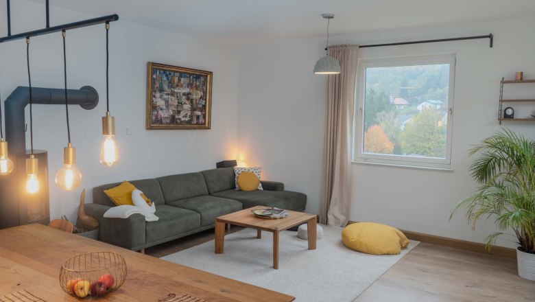 Modernes Wohnzimmer mit grauem Sofa, Holztisch, gro&szlig;en Fenstern und H&auml;ngelampen.