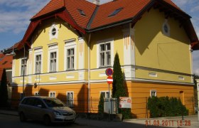 Gelbes und oranges Gebäude mit rotem Dach und geparktem Auto davor.