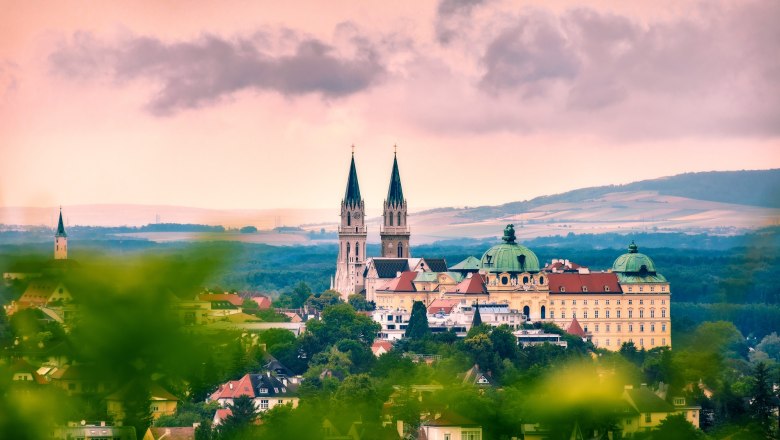 Panoramic view of Stift Klosterneuburg with two church towers and baroque building, surrounded by green landscape and under a cloudy sky.